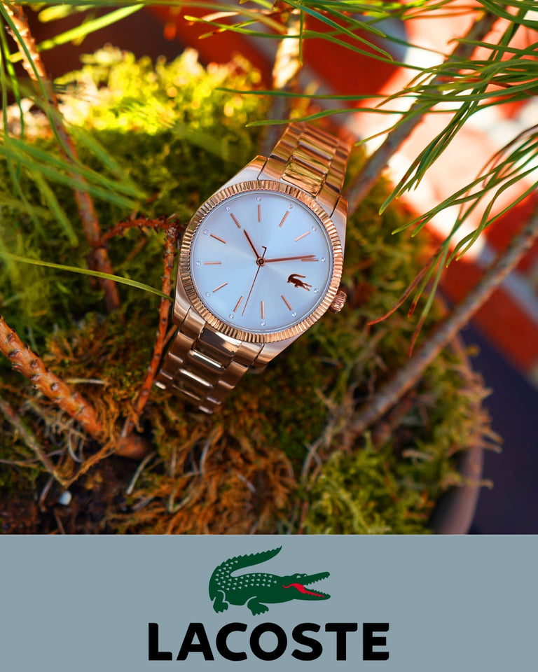 Lacoste ur i guld med hvid skive, forsidevisning, placeret blandt grønne grene og mos